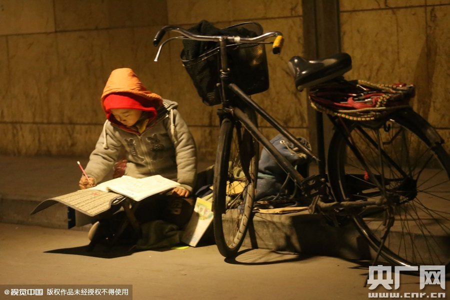 鄭州中原路京廣鐵路大橋下涵洞的馬路牙子邊，一位戴著紅色辣椒帽的小男孩趴在一個(gè)小馬扎上，上面鋪著一張車墊，在微弱的燈光下寫作業(yè)